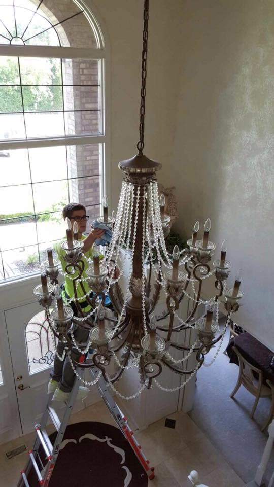 A Glass Guru technician on a tall ladder wiping a bronze chandelier hung with crystal strands in a two-story foyer
