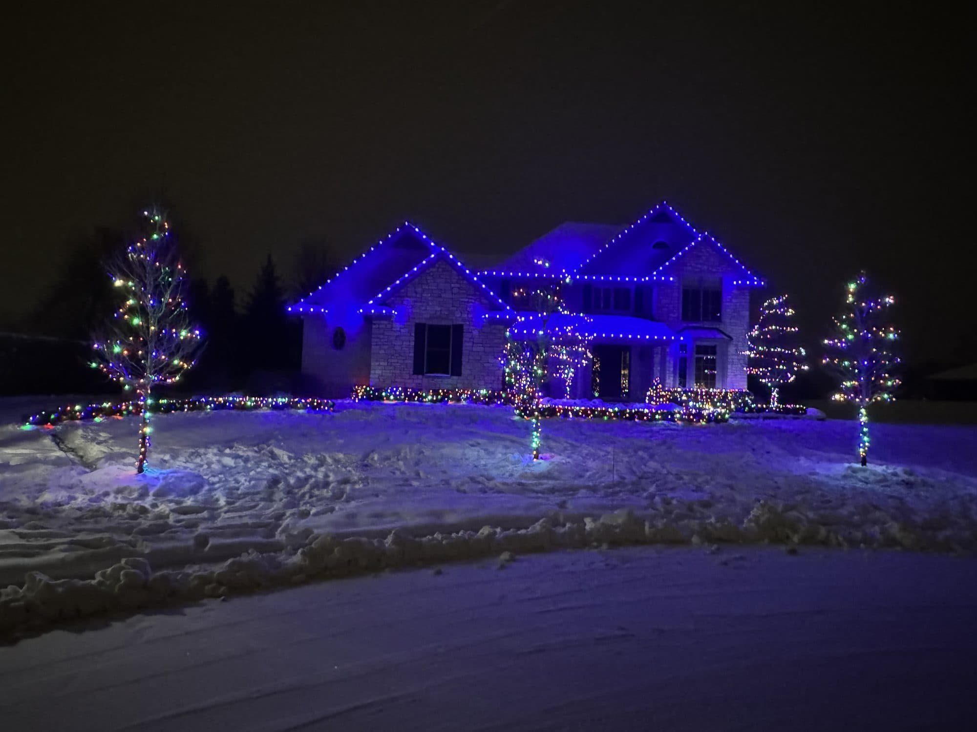 A large home in the snow with blue bulbs on every roofline and trees in the yard wrapped in multicolor lights