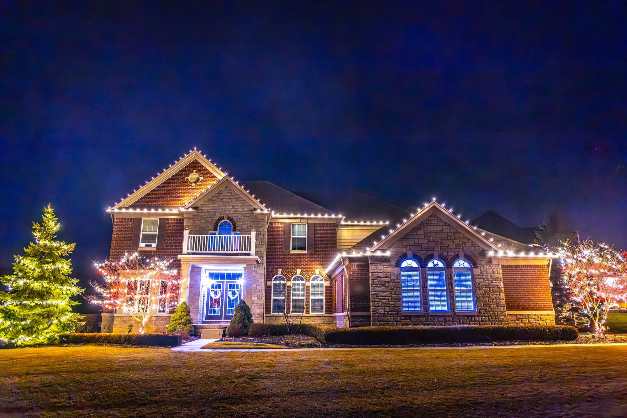 A brick two-story home at night, white bulbs on every roofline, a spruce wrapped in warm white and two trees in red and white