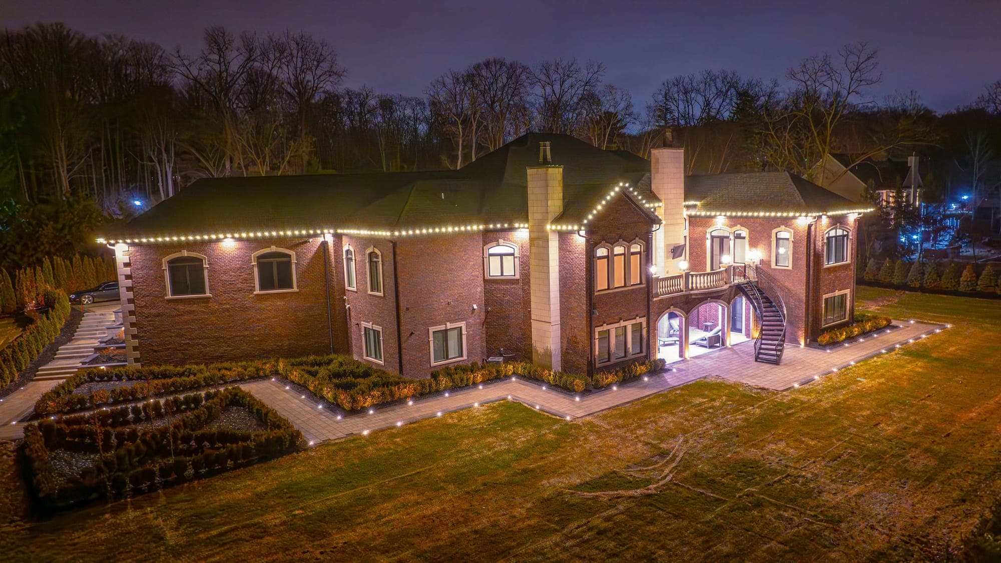 The back of a large brick home from above at night, warm white bulbs on the eaves and gable and a curved stair to the patio