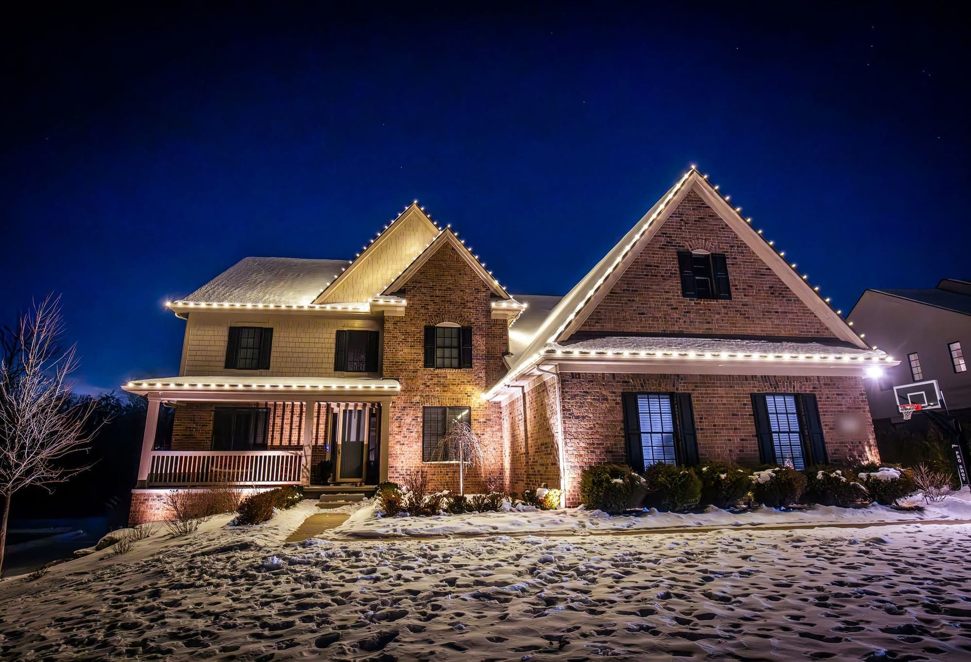 A brick and siding two-story home in the snow, white bulbs outlining each gable and the porch roof