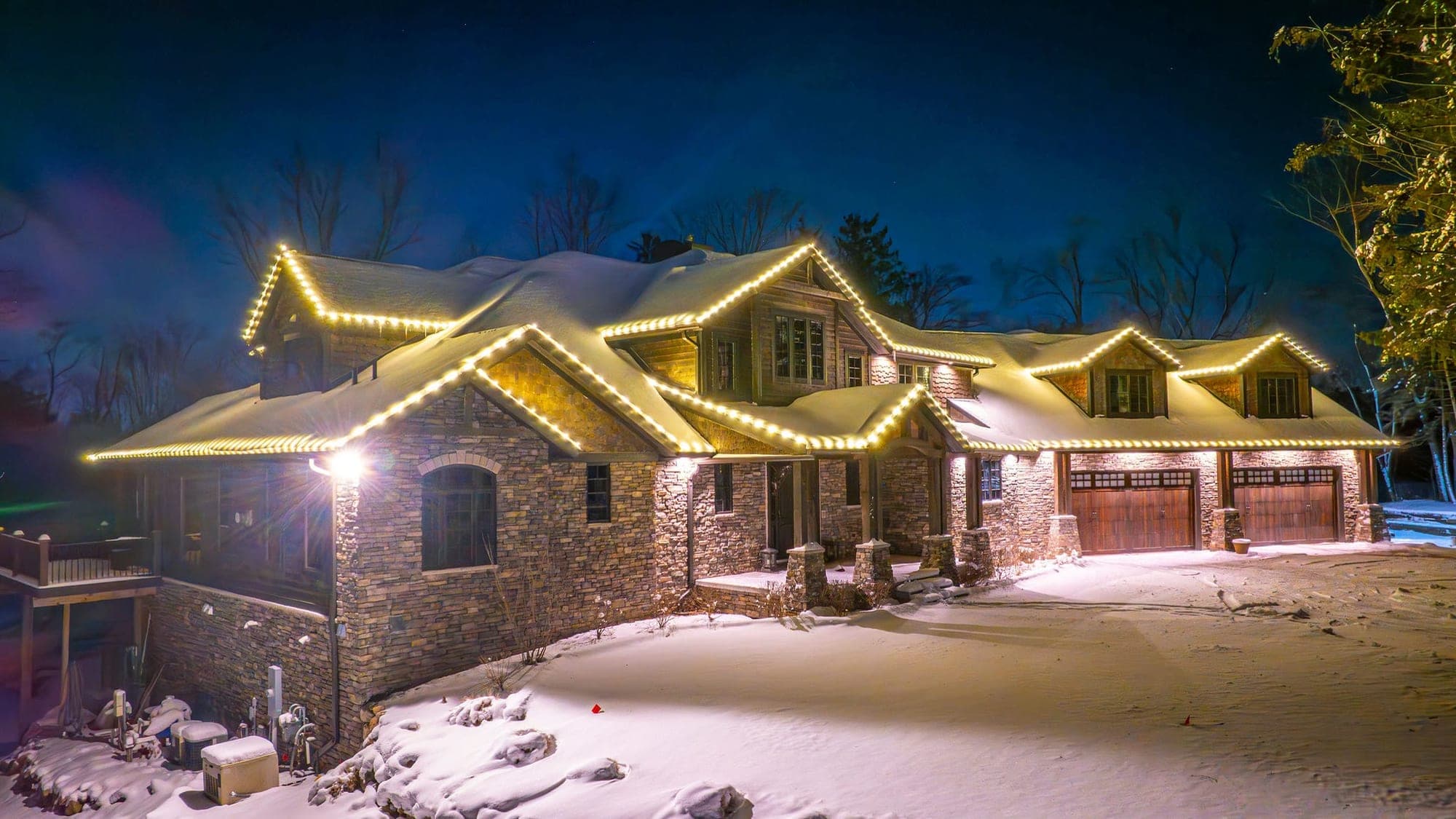 A stone and cedar-shake house under heavy snow at night, warm white bulbs outlining every gable, dormer and eave, with two wooden garage doors at the right