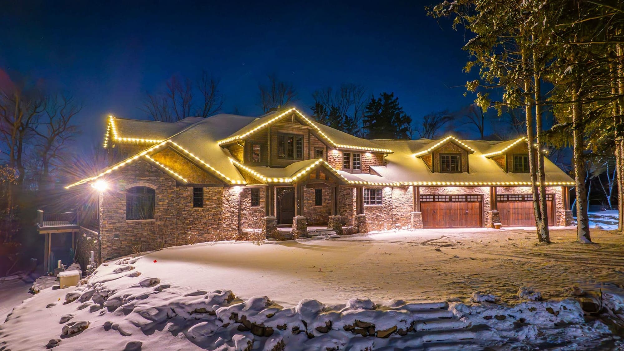 A stone and cedar-shake home under fresh snow at dusk, warm white bulbs outlining every gable, dormer and eave