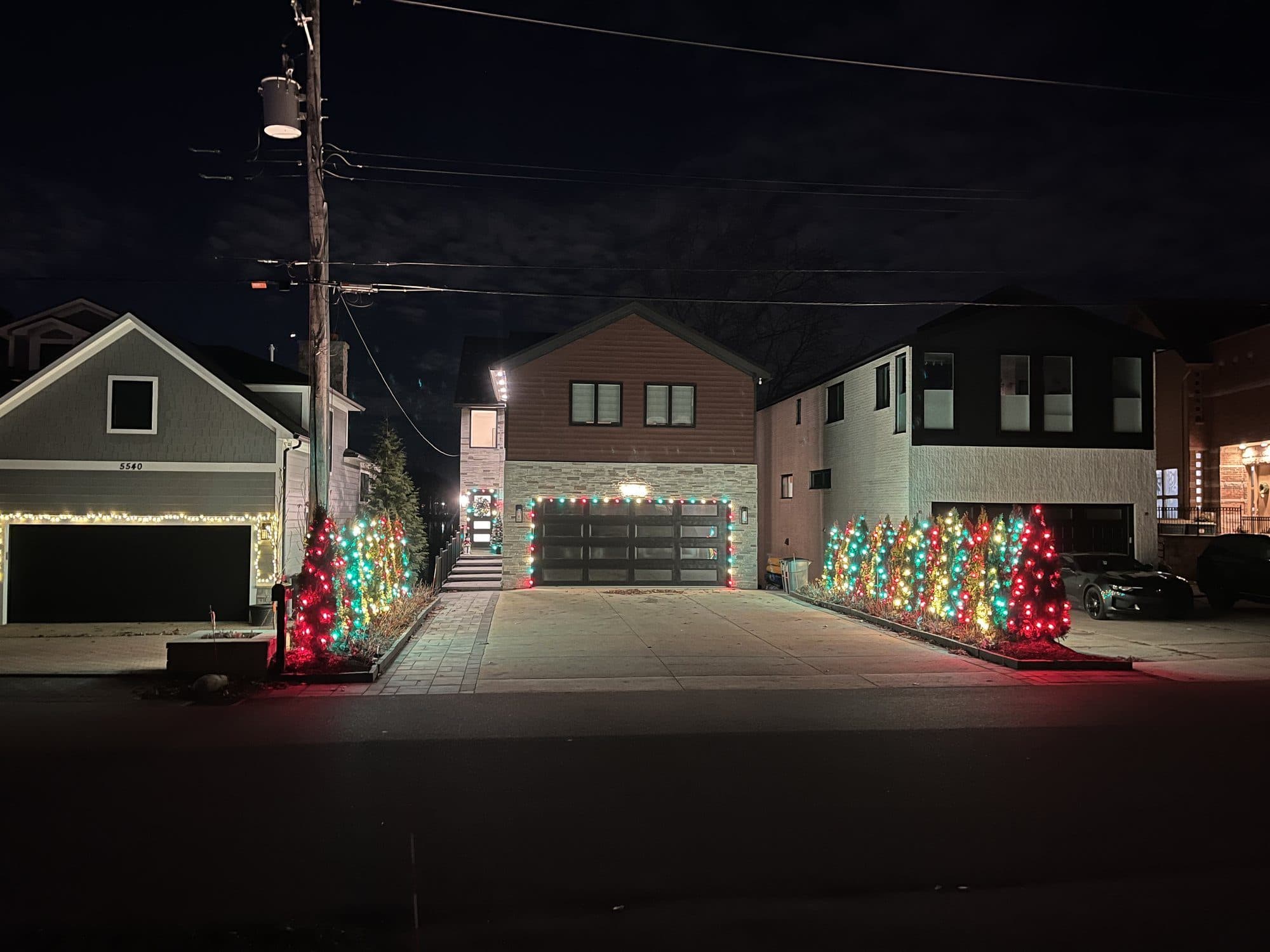 Three new homes side by side at night, the driveways lined with rows of small trees lit in red, green and white