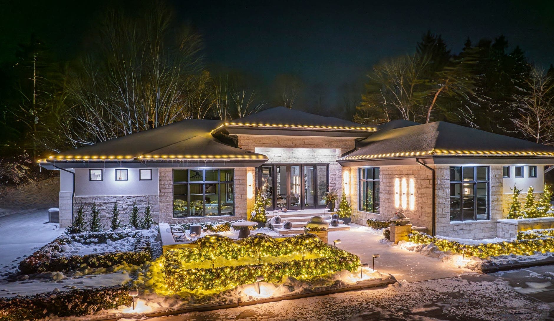 A modern single-story home at night, warm white bulbs on the eaves, net lights on the hedges and lit evergreens by the entry
