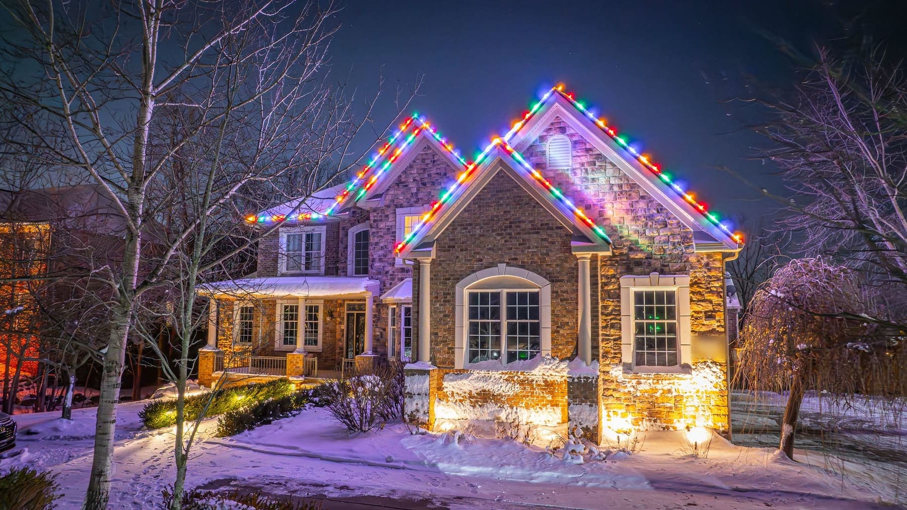 A stone and brick home in the snow at night, multicolor bulbs tracing its two front gables