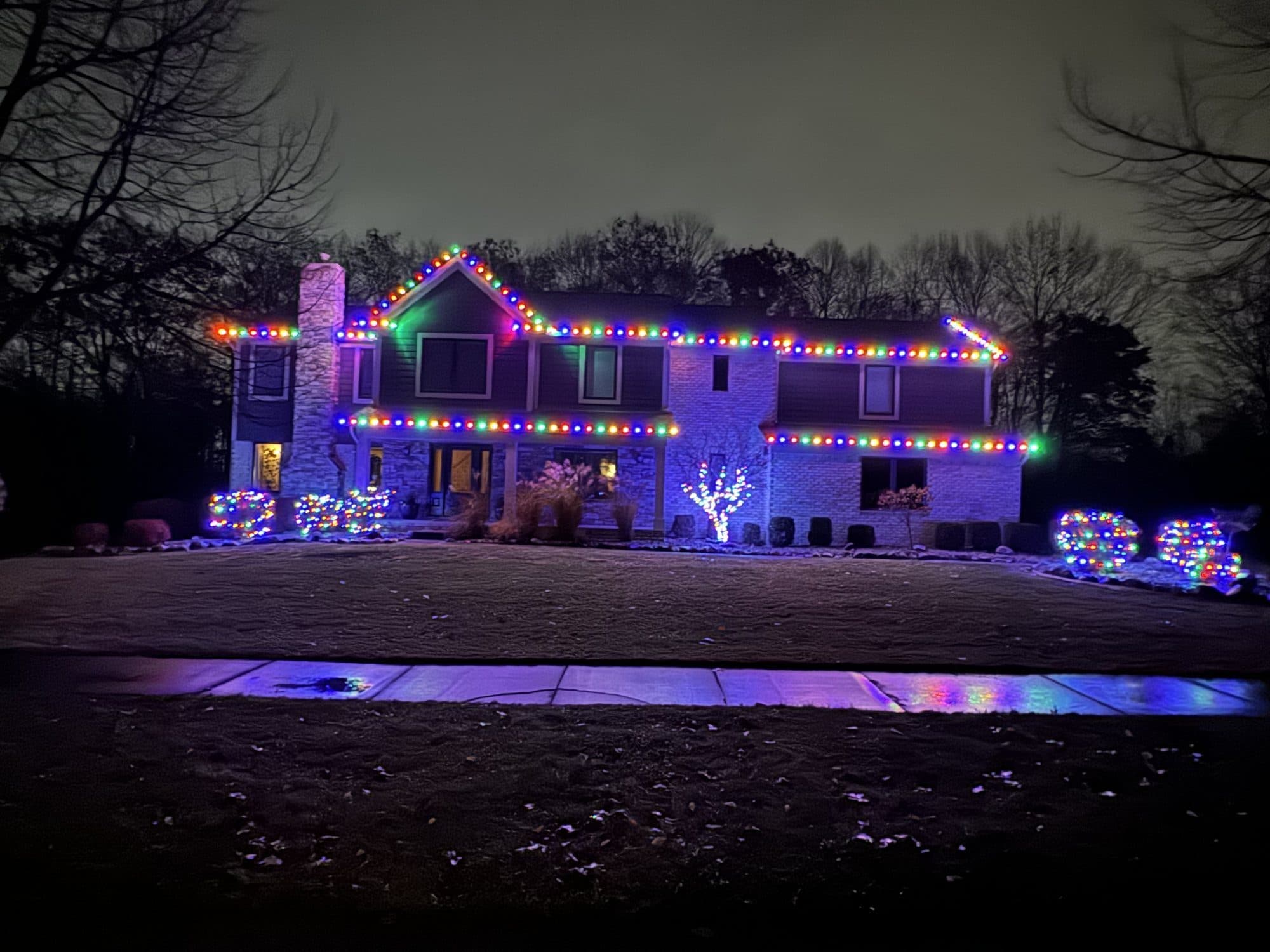 A two-story home at night with multicolor bulbs on every roofline and lit shrubs along the front