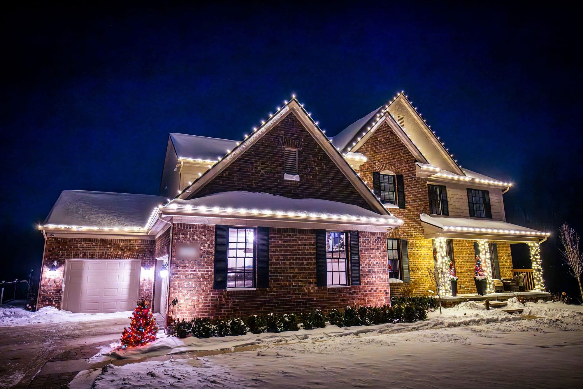 A red brick two-story home in the snow, white bulbs along every roofline and the porch columns wrapped in lights