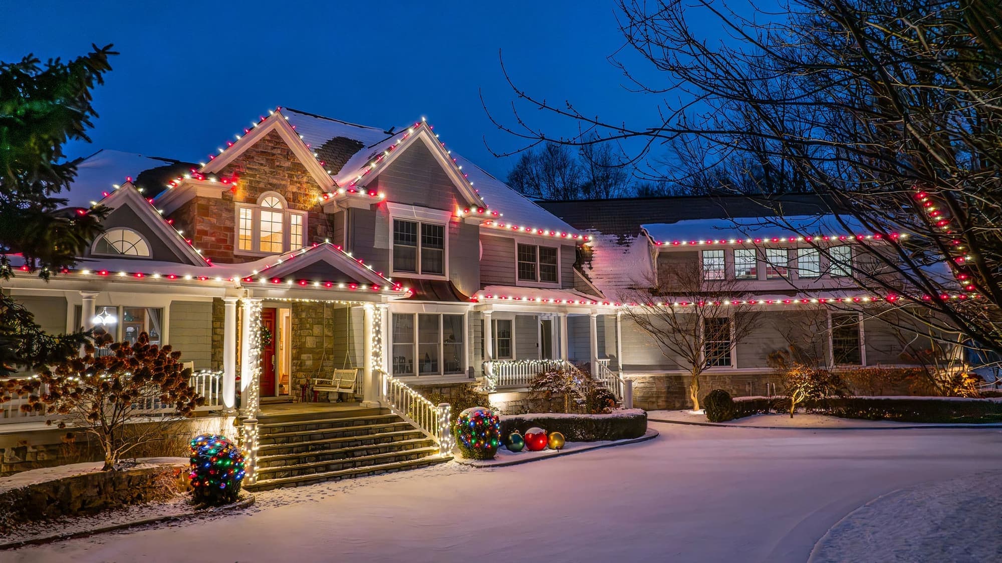 A shingle-sided home in the snow, red and white bulbs on every roofline, white lights on the porch columns and stair railings