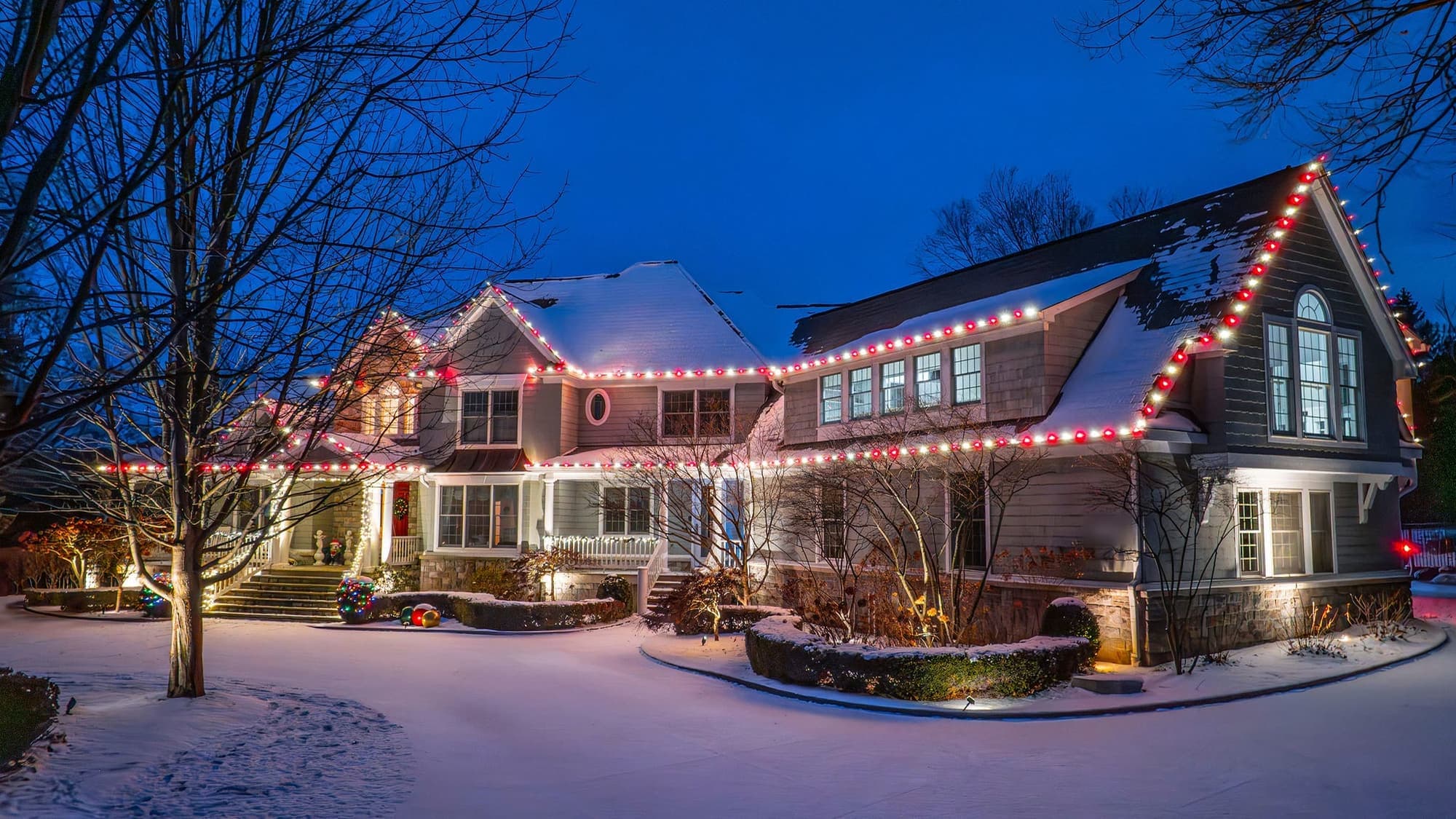 A large shingle-sided home at blue hour after a snowfall, red and white bulbs alternating along every roofline and gable