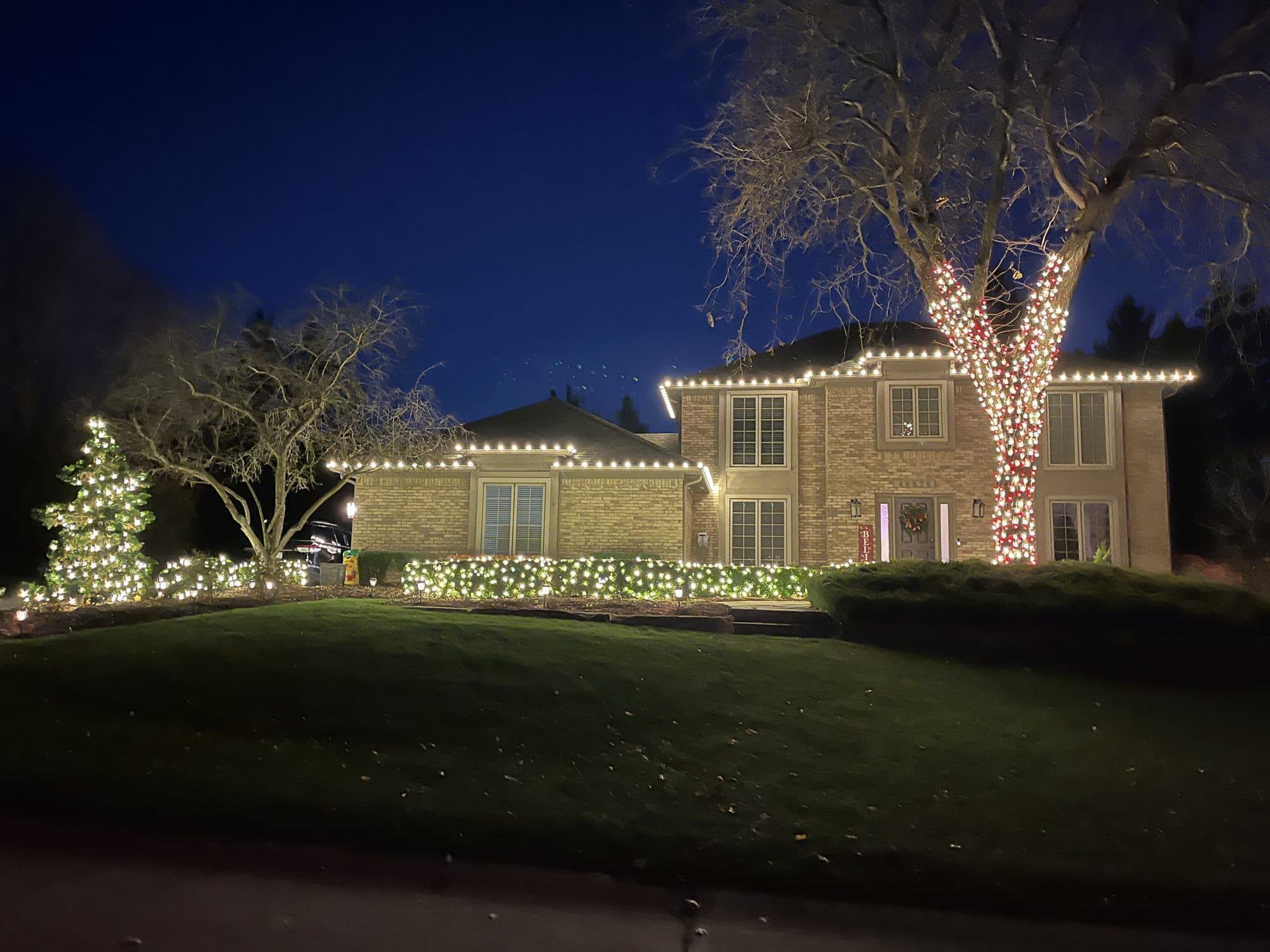 A two-story home with warm white rooflines, a large tree wrapped in red and white lights and a lit evergreen