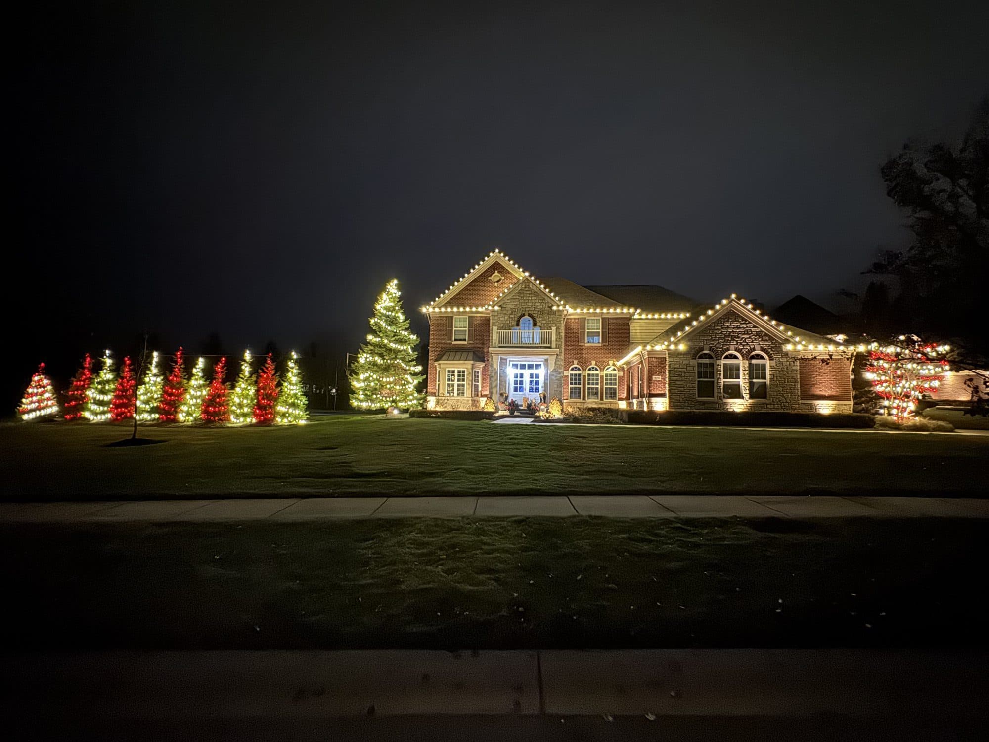 A brick home with warm white rooflines and a row of small trees across the lawn lit in red, green and white