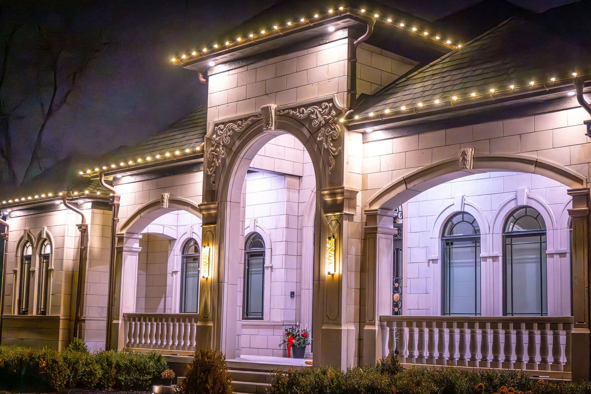 The carved arched entry of a limestone house at night, warm white bulbs along the eaves above it