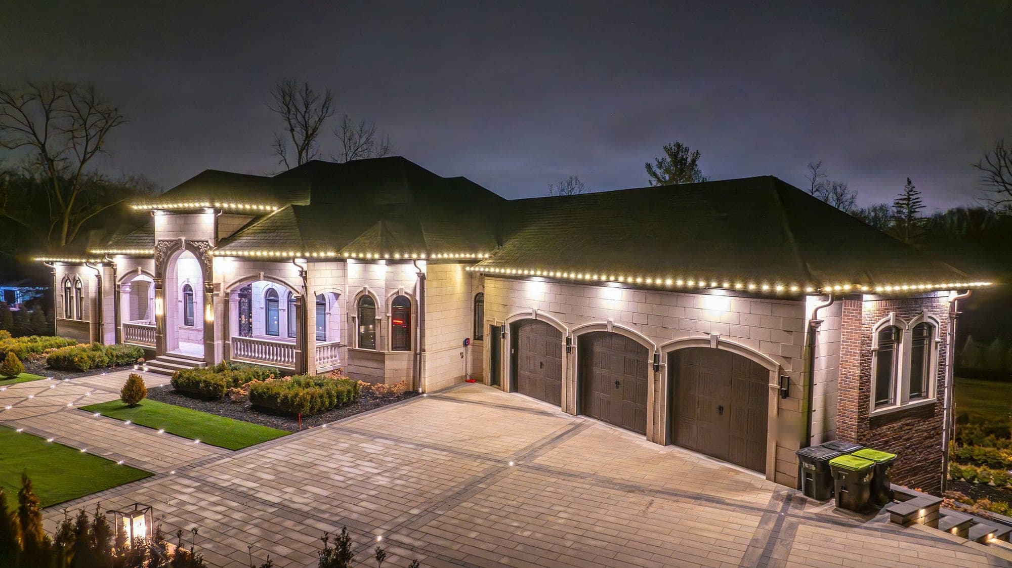 A limestone house with a three-car garage seen from above at night, warm white bulbs along every eave