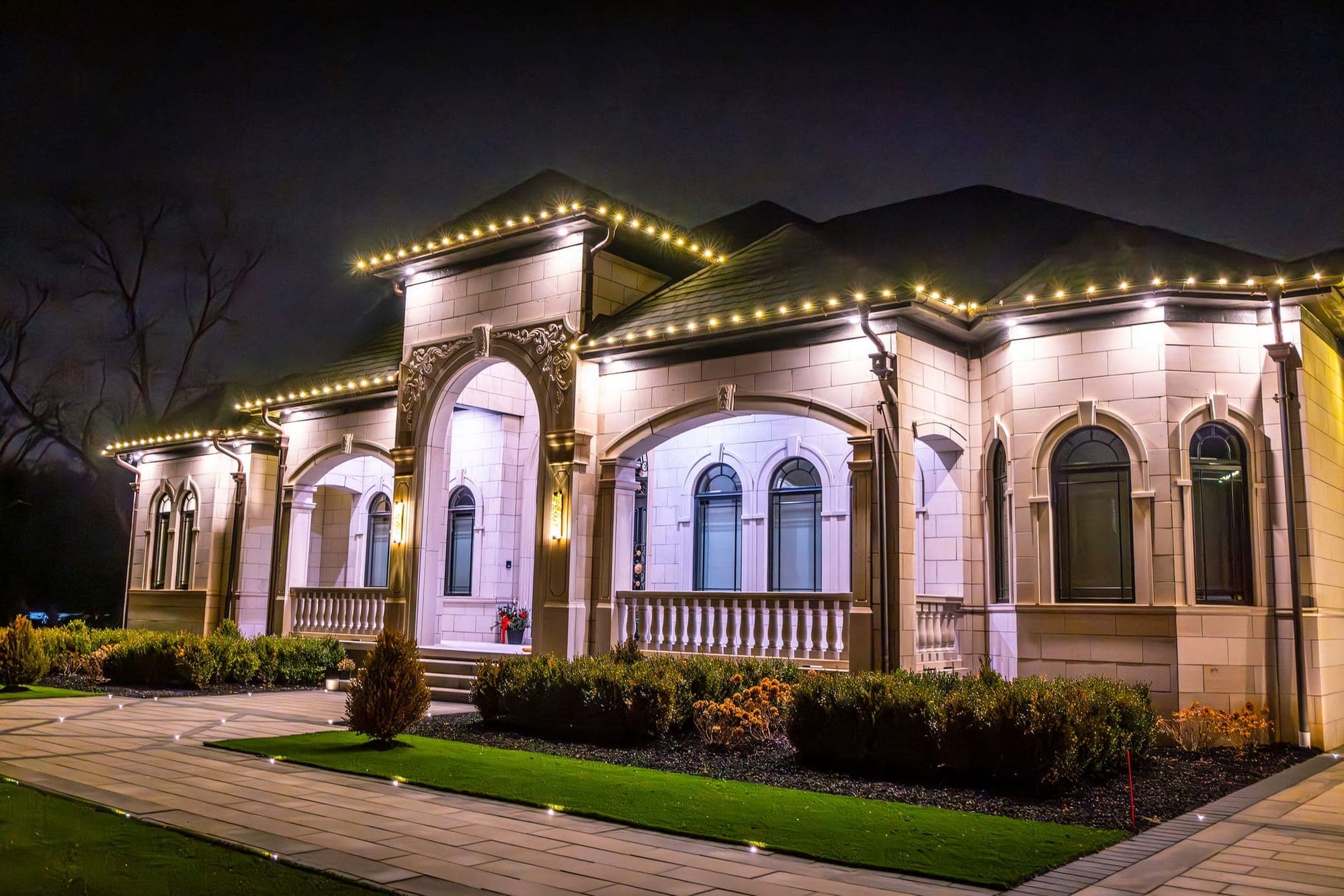 A limestone house with arched porches at night, warm white bulbs running the full length of every eave