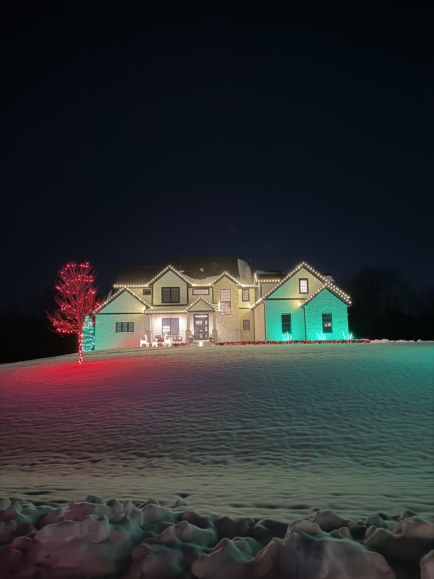 A two-story home in the snow with warm white rooflines, a wall washed in teal light and a tree wrapped in red