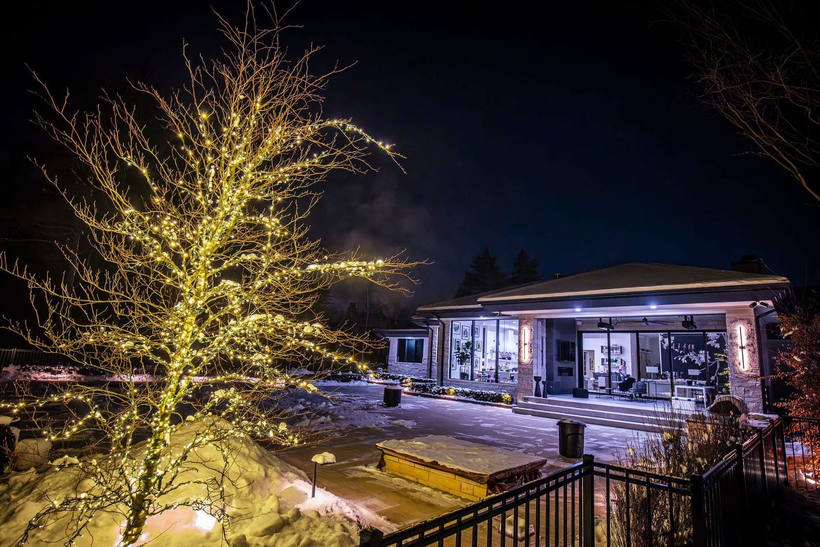 A bare tree wrapped branch by branch in warm white lights beside the snowy patio of a modern home