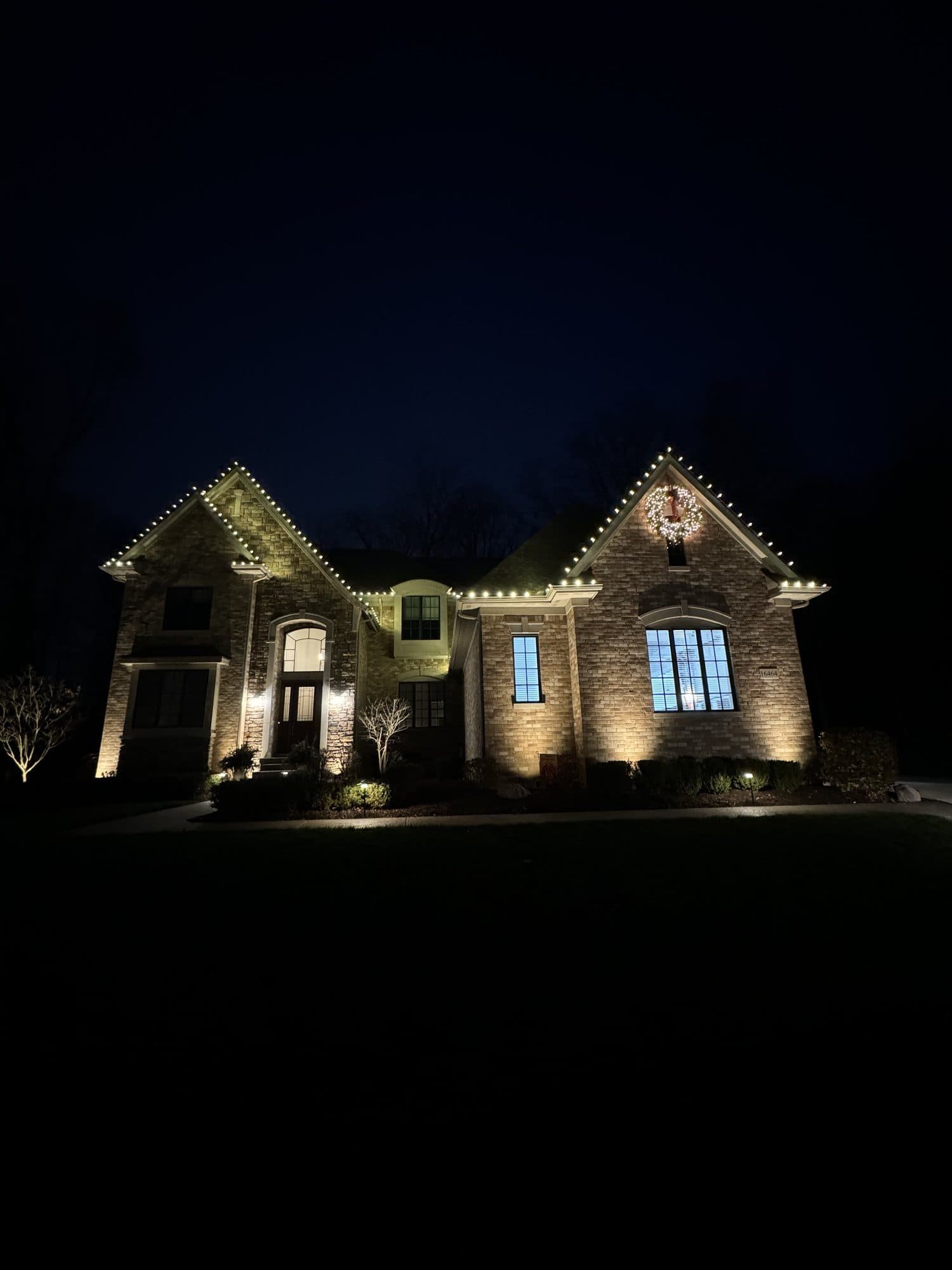 A brick home at night with warm white bulbs along its gables and eaves and a lit wreath on the front gable
