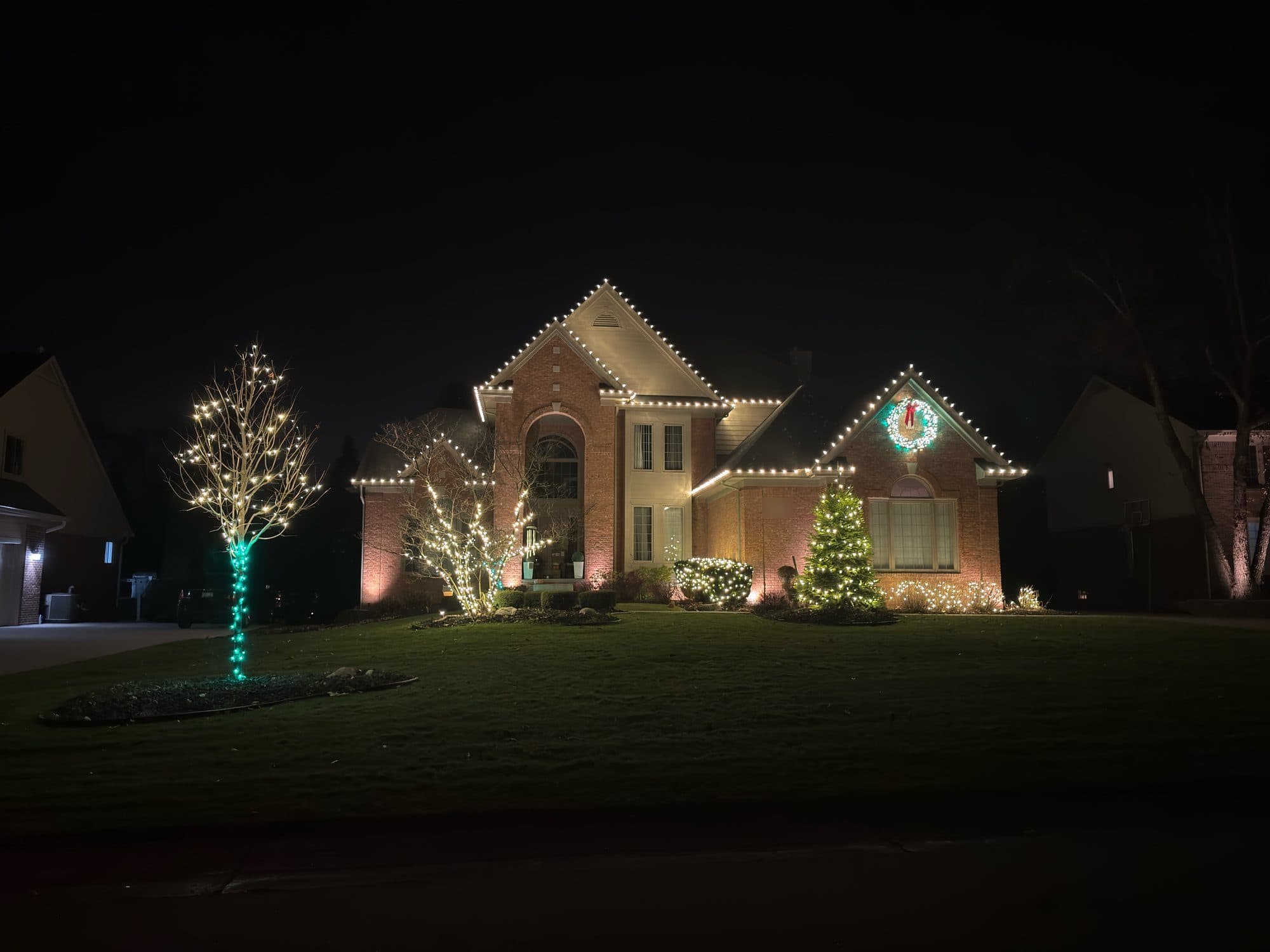 A brick home at night with warm white bulbs on every gable, a lit wreath on the right gable, a tree with a teal-wrapped trunk and a lit spruce by the door