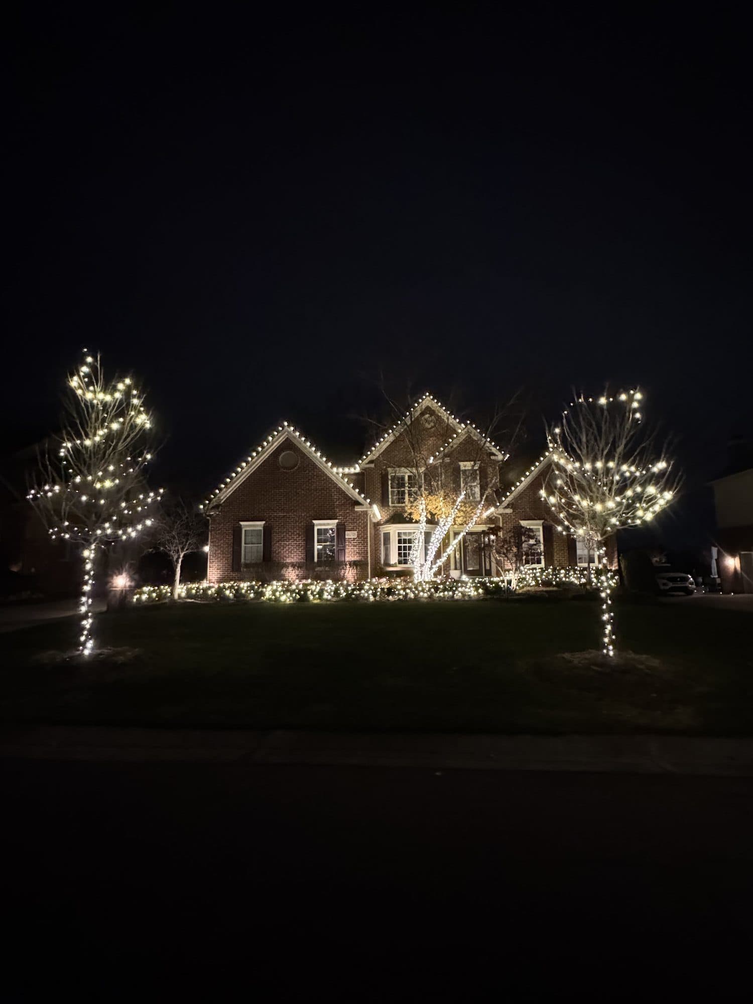 A brick home with warm white bulbs on its gables and two front-yard trees wrapped in white lights