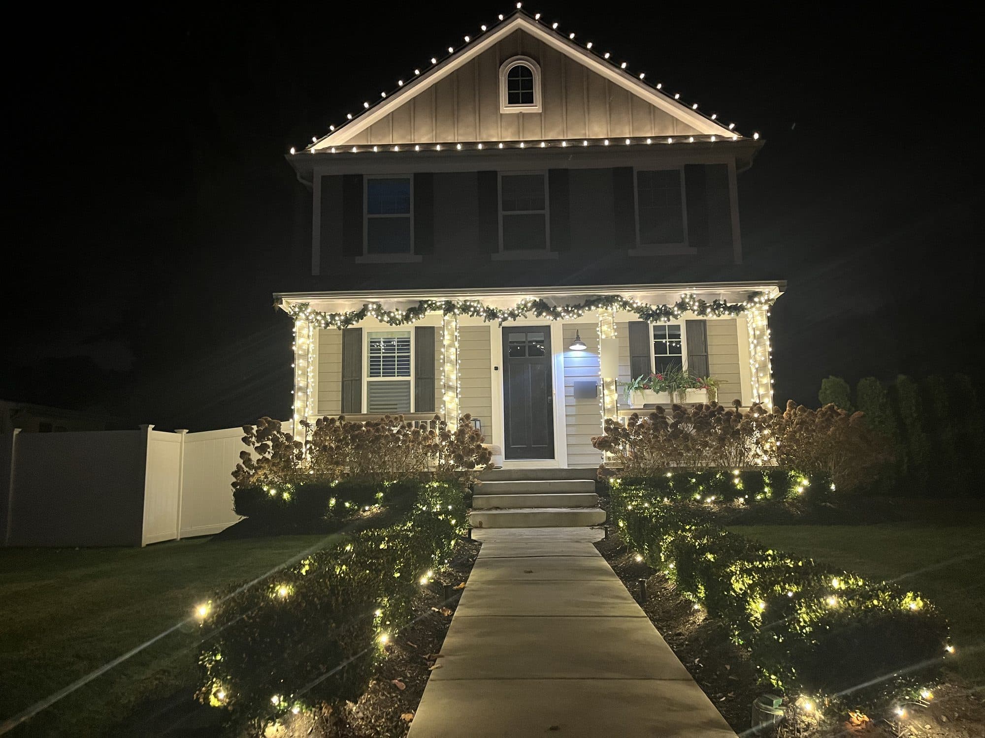 A two-story home with warm white bulbs along the roofline, lit porch columns and white lights lining the front walk