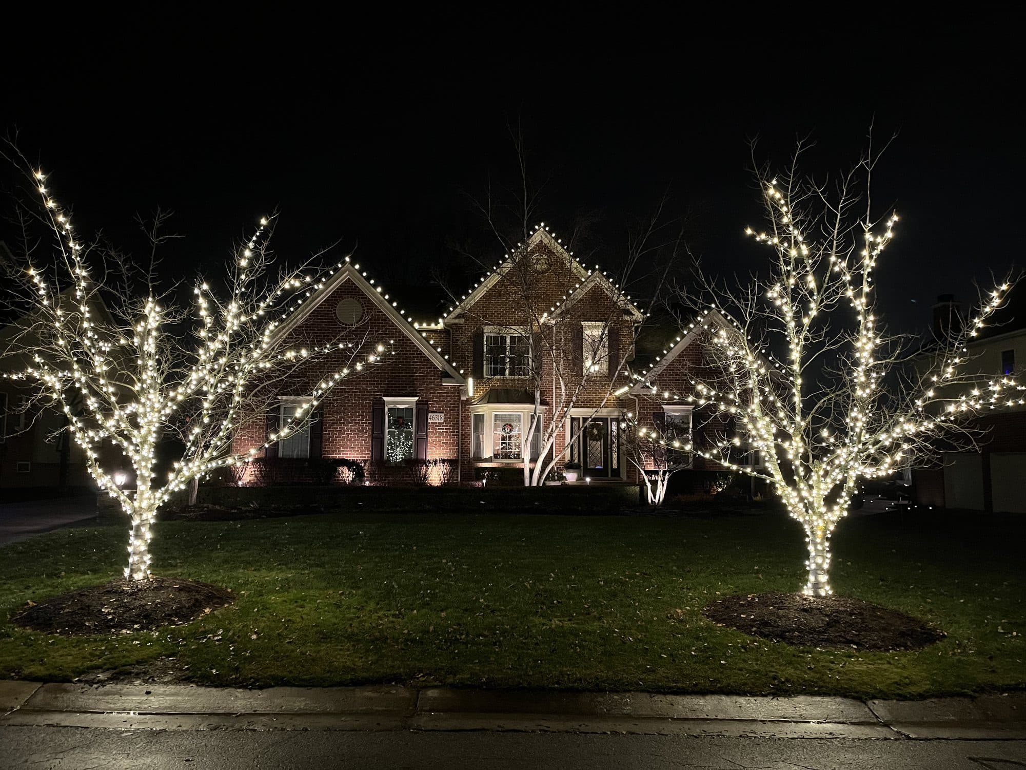 A brick home at night with two front-yard trees wrapped in white lights from trunk to branch tips