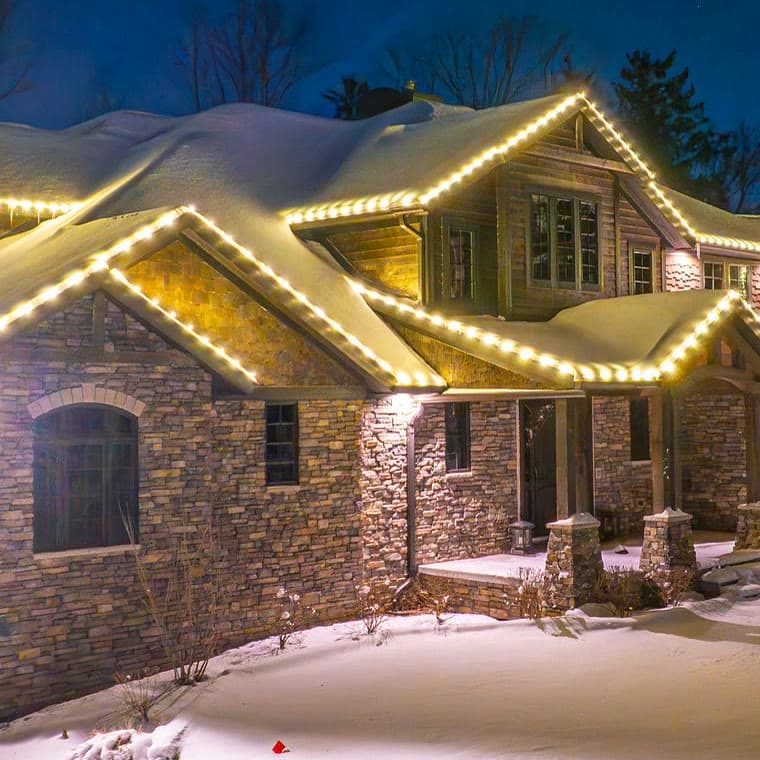 Warm white bulbs following the stacked gables and a dormer of a stone and cedar-shake home under snow