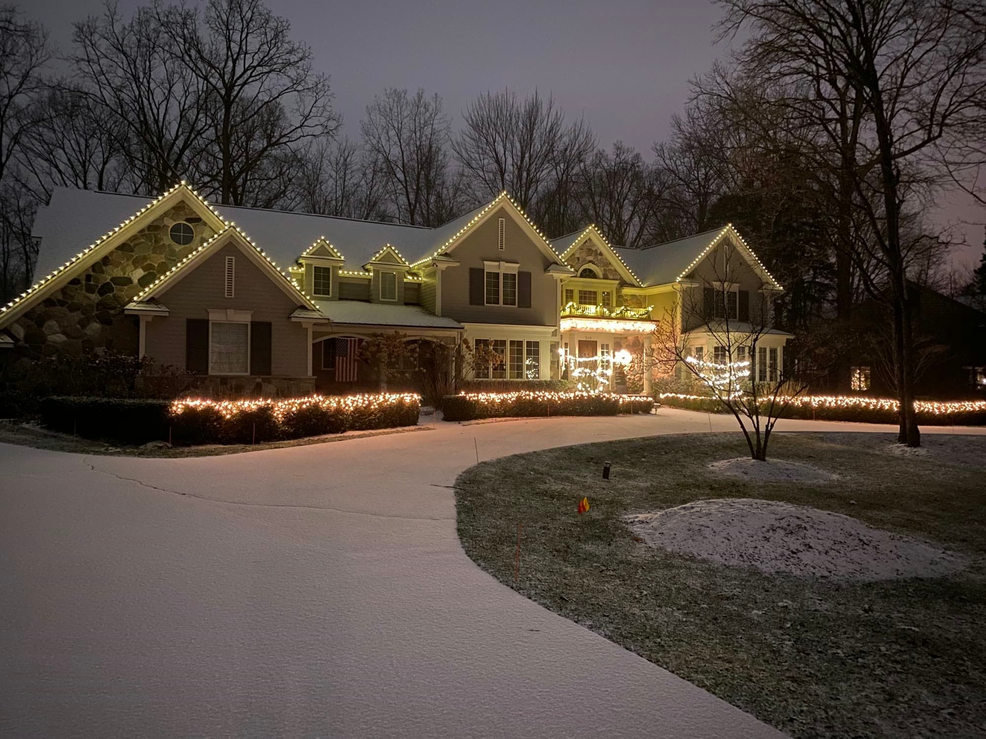 A large home at dusk after a light snow, warm white lights along every gable and eave and across the front hedges
