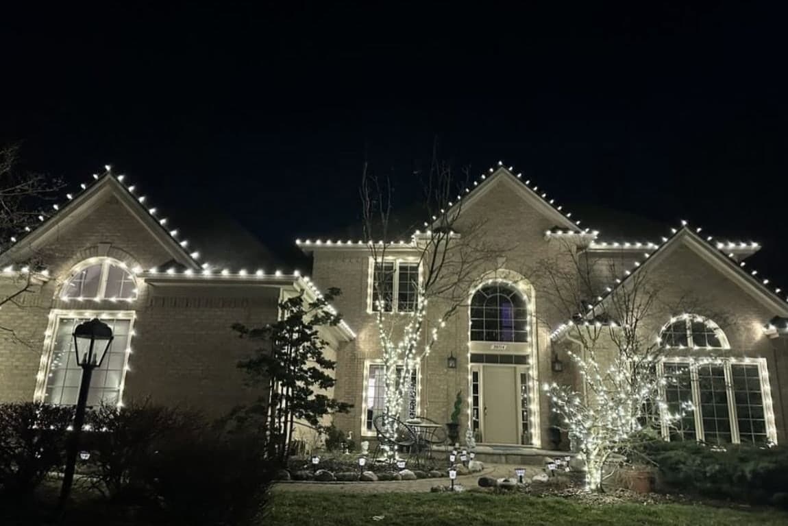 A two-story home at night with warm white bulbs outlining every roof edge and lights wrapped through the front trees