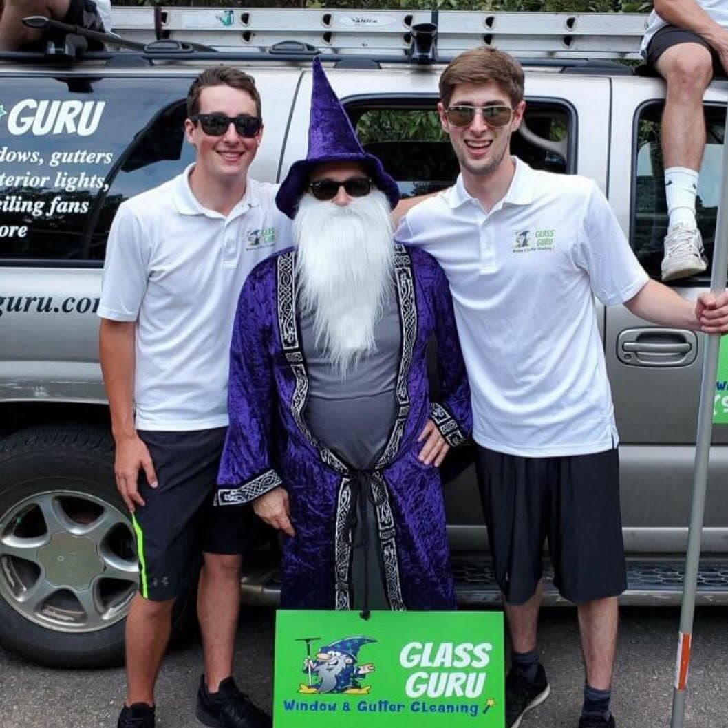 Two people in Glass Guru polo shirts standing either side of the wizard mascot in costume, in front of a silver SUV lettered with the Glass Guru name