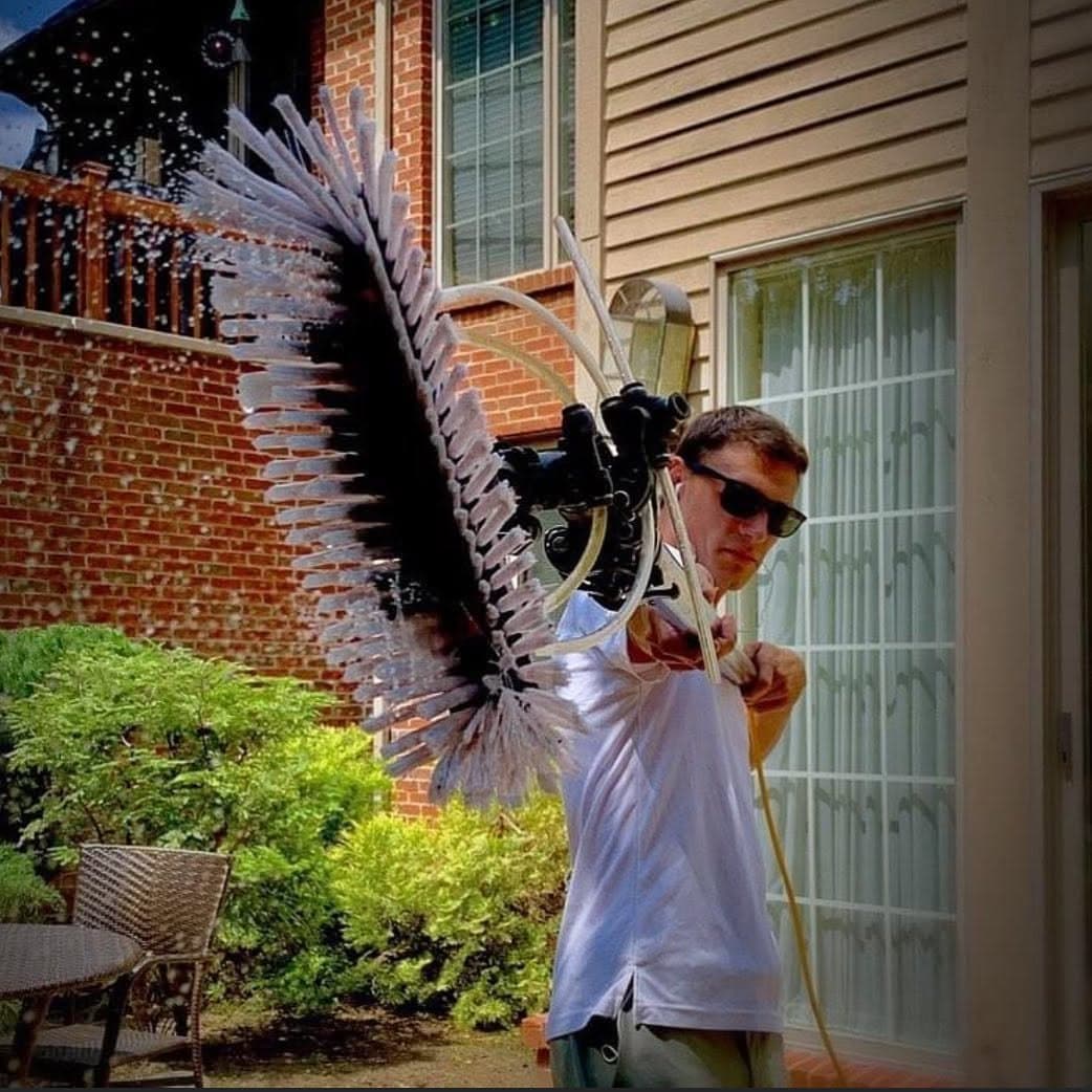 A technician holds the brush head of a water-fed pole toward the camera, water spraying off the bristles, beside a brick and sided house