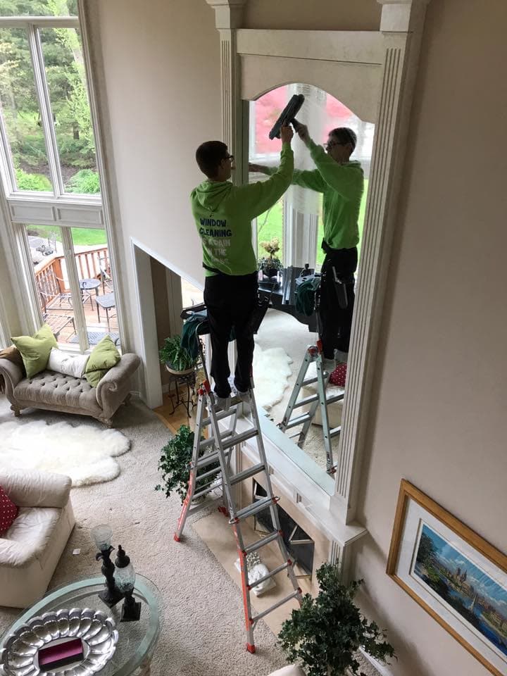 A Glass Guru technician on a stepladder cleaning a tall arched mirror above a fireplace in a two-story living room