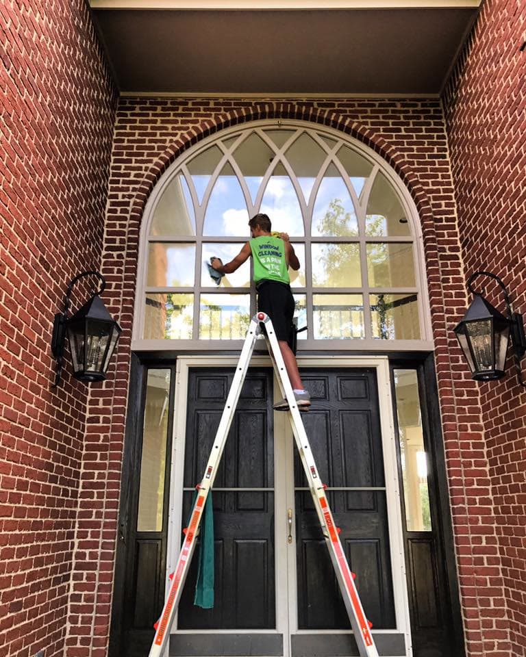 A Glass Guru technician on a stepladder cleaning a tall arched transom above a pair of front doors in a brick entry