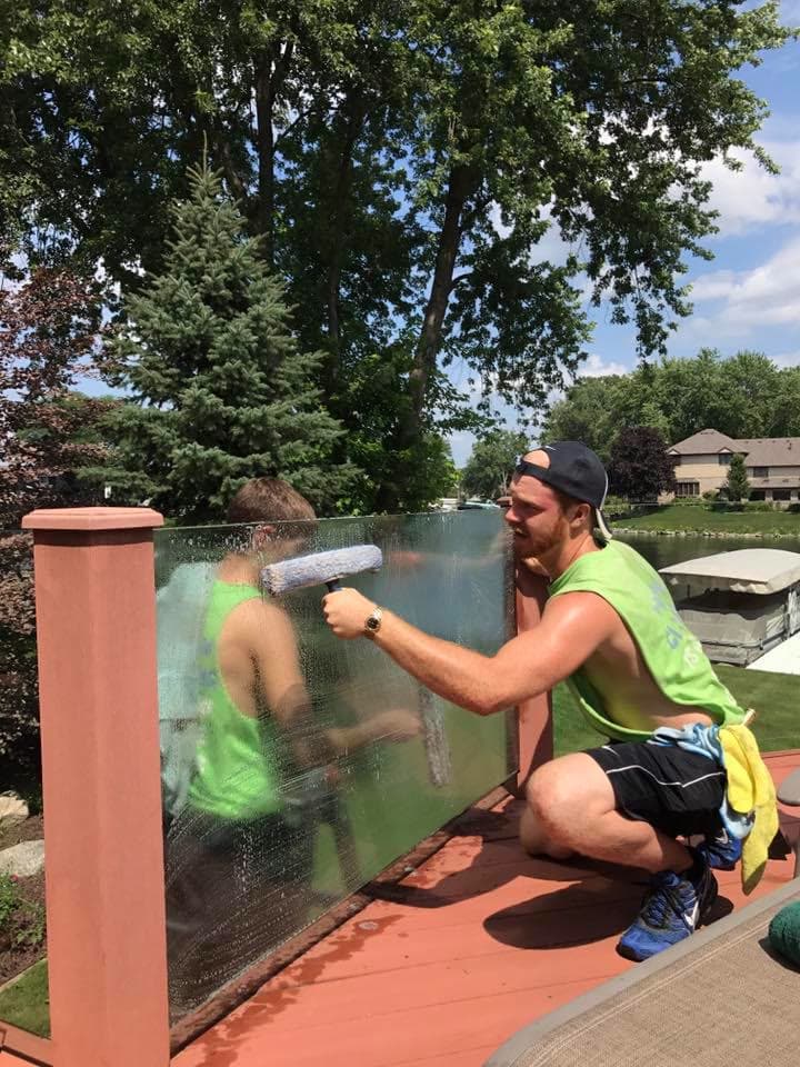 Two Glass Guru technicians washing the glass panels of a deck railing, water and a covered boat lift behind them