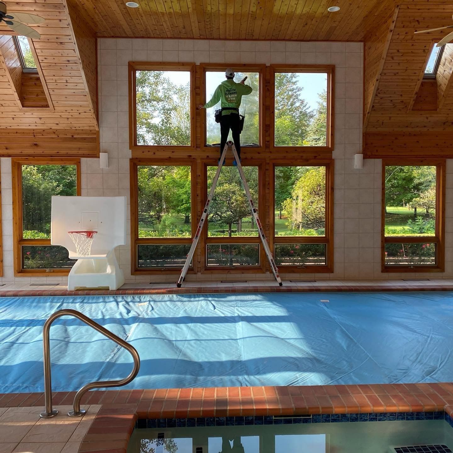 A Glass Guru technician on a stepladder cleaning a wood-framed window wall above an indoor pool