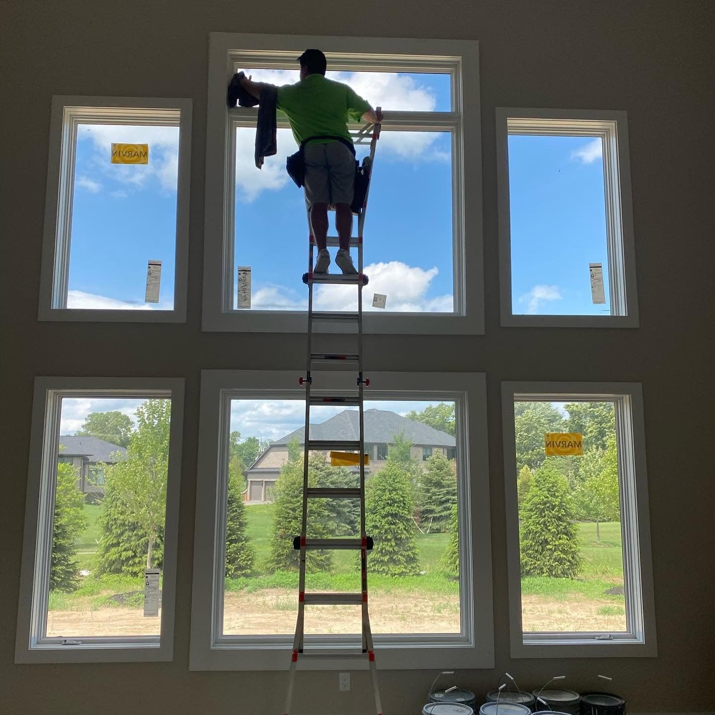 A Glass Guru technician on a ladder cleaning the inside face of a tall window wall in a two-story room